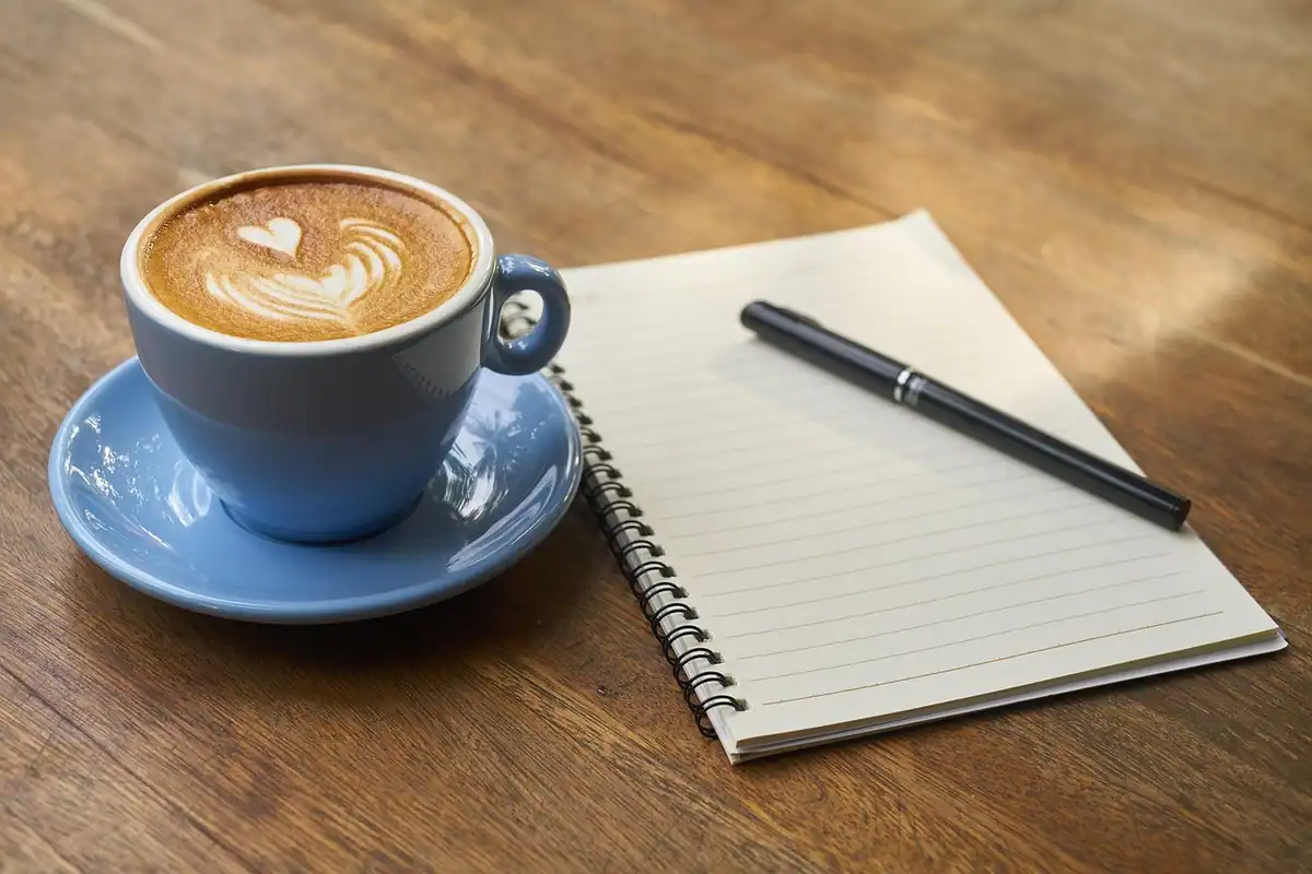 Caiet cu spirală, pix și cafea cu spumă pe un blat de lemn, pentru notițe scurte