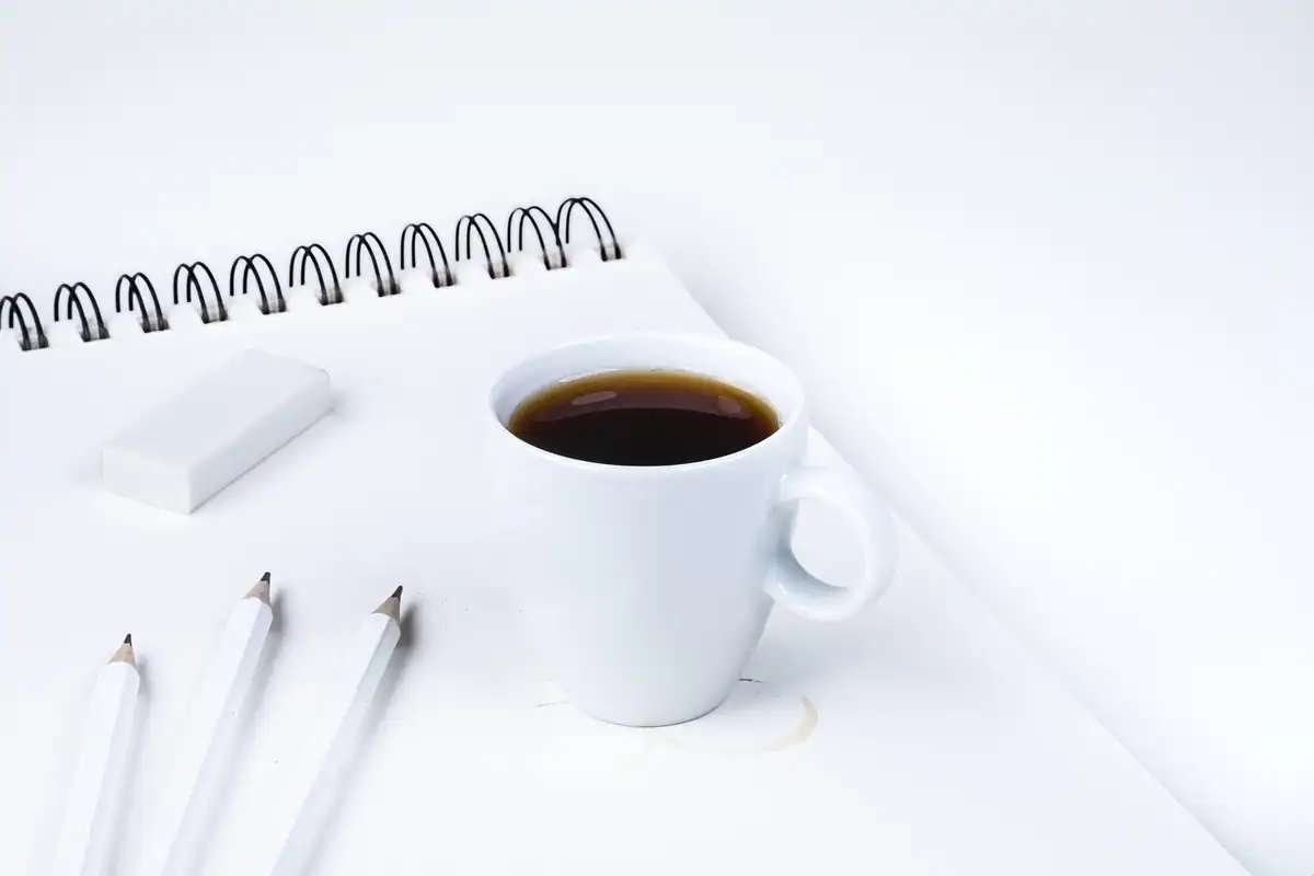 Cană cu cafea neagră lângă un caiet cu spirală și creioane, pe un birou alb