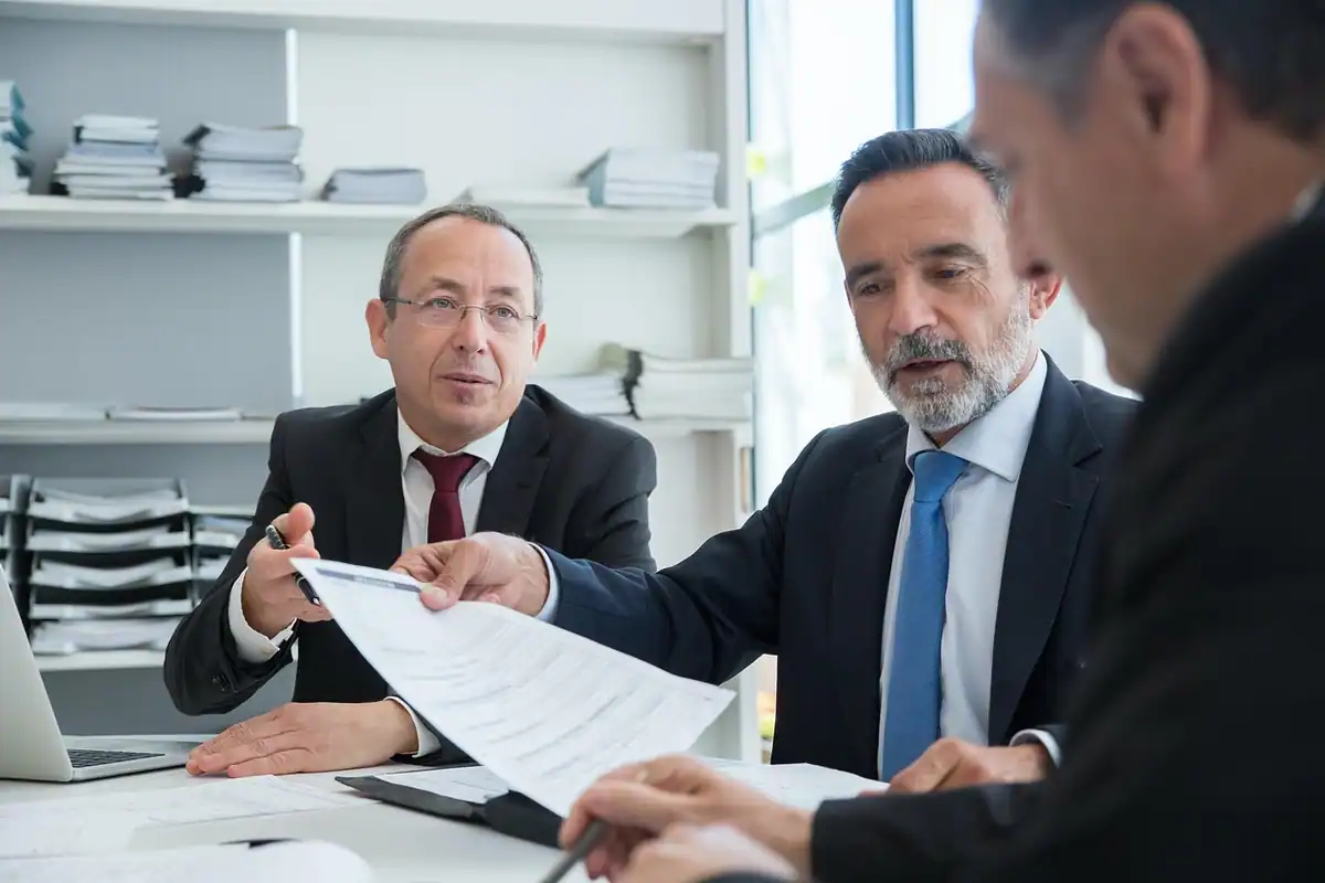 Doi bărbați în costum discută un document întins pe masă, la o întâlnire de lucru