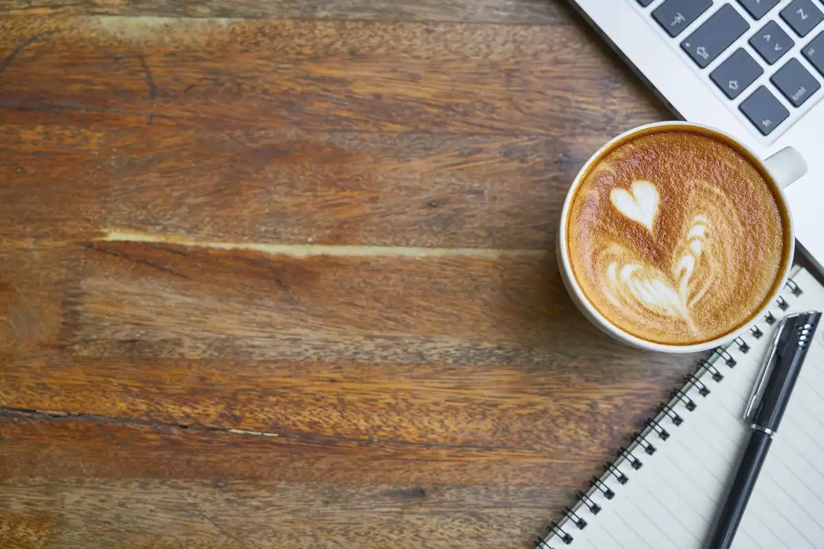 Cafea cu spumă în formă de inimă, lângă laptop și caiet, pe un birou din lemn