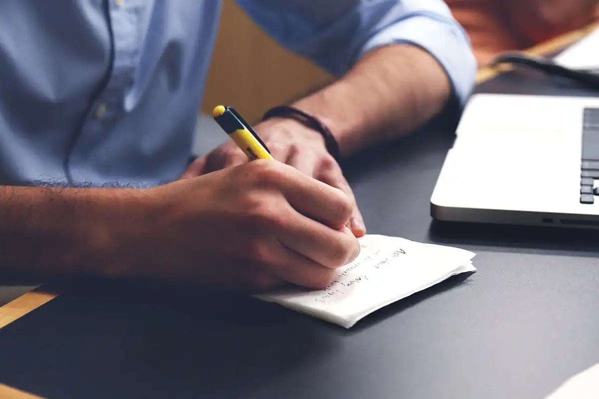 Mână care scrie cu un marker galben într-un carnet, lângă un laptop, la birou