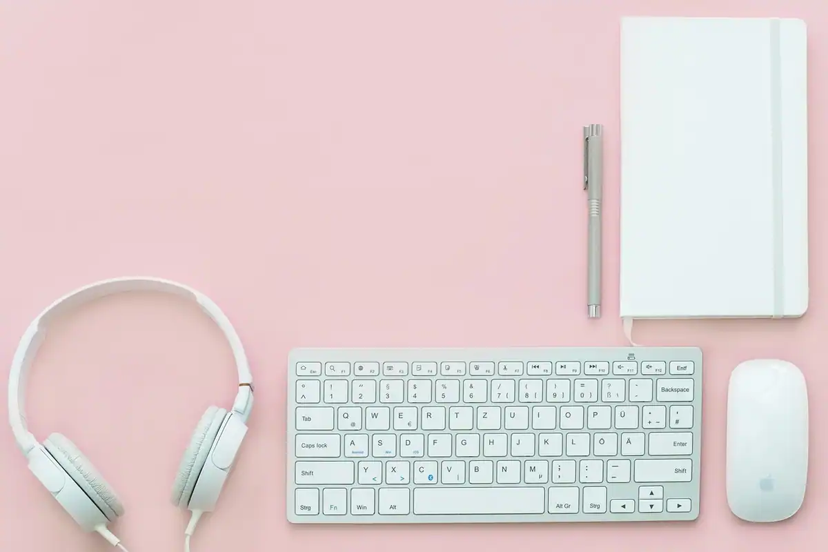 Birou ordonat, cu cască audio, tastatură, mouse și caiet, pe un fundal roz