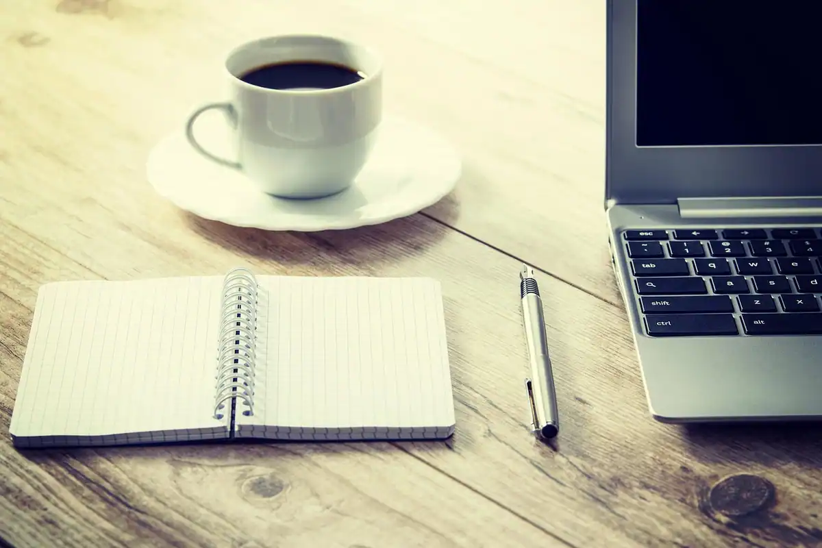 Caiet deschis, cană cu cafea și laptop pe o masă de lemn, pentru stabilirea obiectivelor
